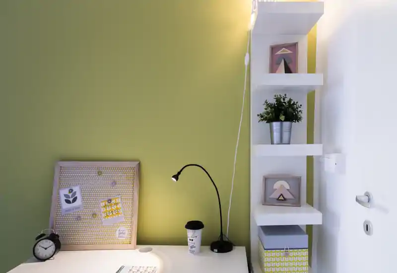 Angolo scrivania ordinato con lampada moderna, bacheca decorativa, pianta in vaso e mensole: un piccolo spazio di lavoro ben curato, ideale per studio o smart working.