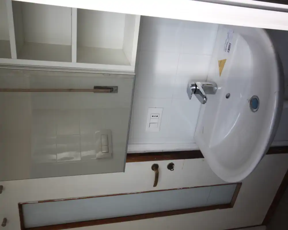 Piccolo lavabo da bagno con miscelatore cromato moderno, specchio con mobiletto e mensole aperte sopra; piastrelle bianche sullo sfondo e porta laterale.
