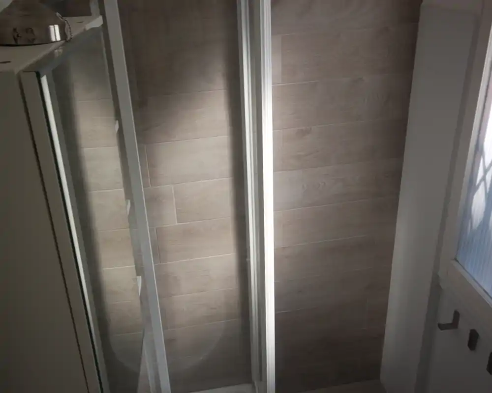 Angolo bagno con cabina doccia e piastrelle effetto legno; illuminazione neutra e disposizione compatta.