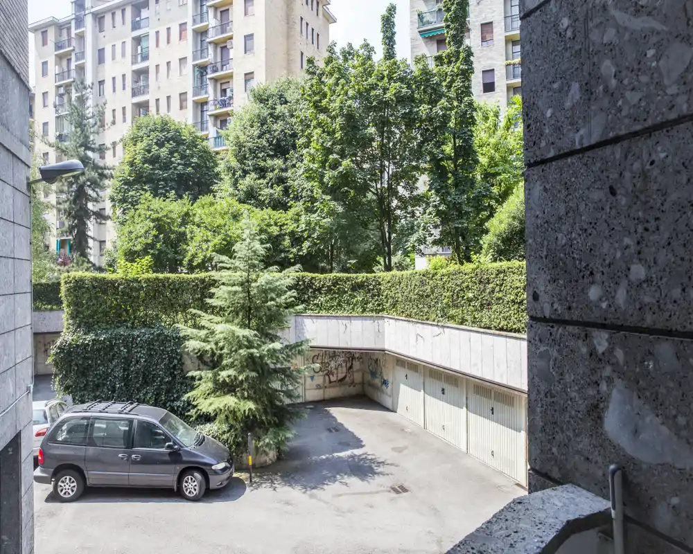 Cortile residenziale soleggiato con parcheggio, box auto e vegetazione matura; adatto a mostrare aree esterne condominiali.