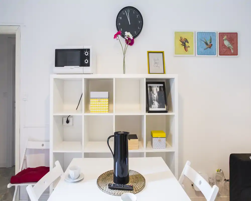 Accogliente zona pranzo moderna e luminosa con tavolo bianco e sedie, mobile a cubi decorativo, caraffa nera e quadri alle pareti — perfetta per annunci di monolocali o bilocali.