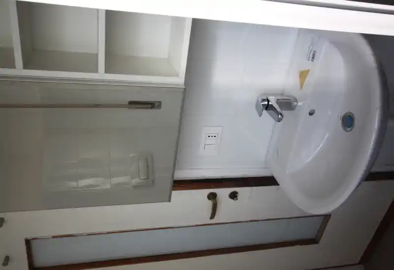 Zona de lavabo de baño compacta con grifo cromado moderno, armario con espejo y estanterías abiertas arriba; alicatado blanco y puerta lateral.