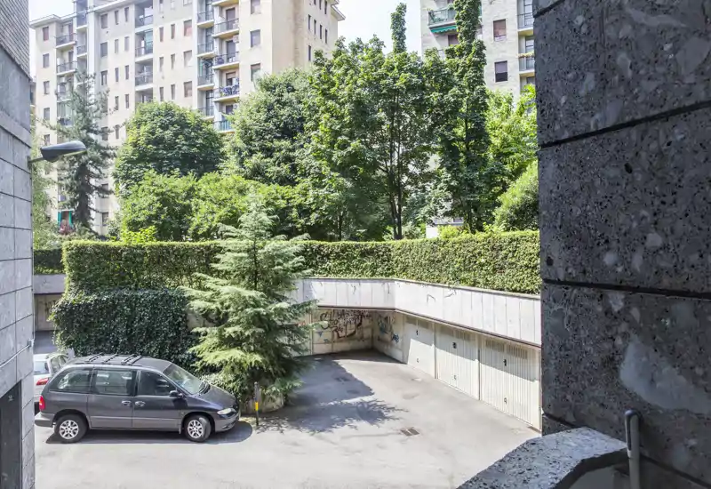 Patio residencial soleado con plazas de aparcamiento, puertas de garaje y vegetación adulta; adecuado para mostrar zonas exteriores comunitarias.