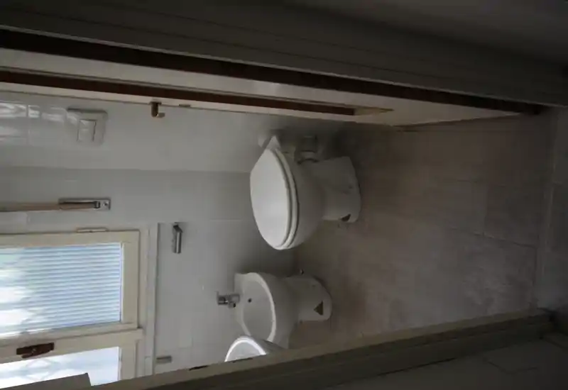 Pequeño baño con inodoro, bidé y lavabo visibles, azulejos neutros y una ventana esmerilada que aporta luz natural.