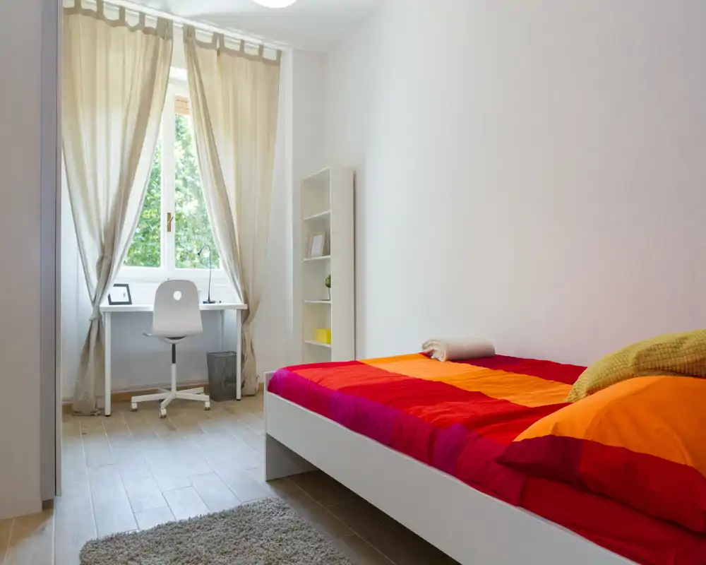 Bright, tidy bedroom with a colorful single bed and a small workspace by the window. Natural light and simple, modern furniture create a welcoming atmosphere ideal for short stays.