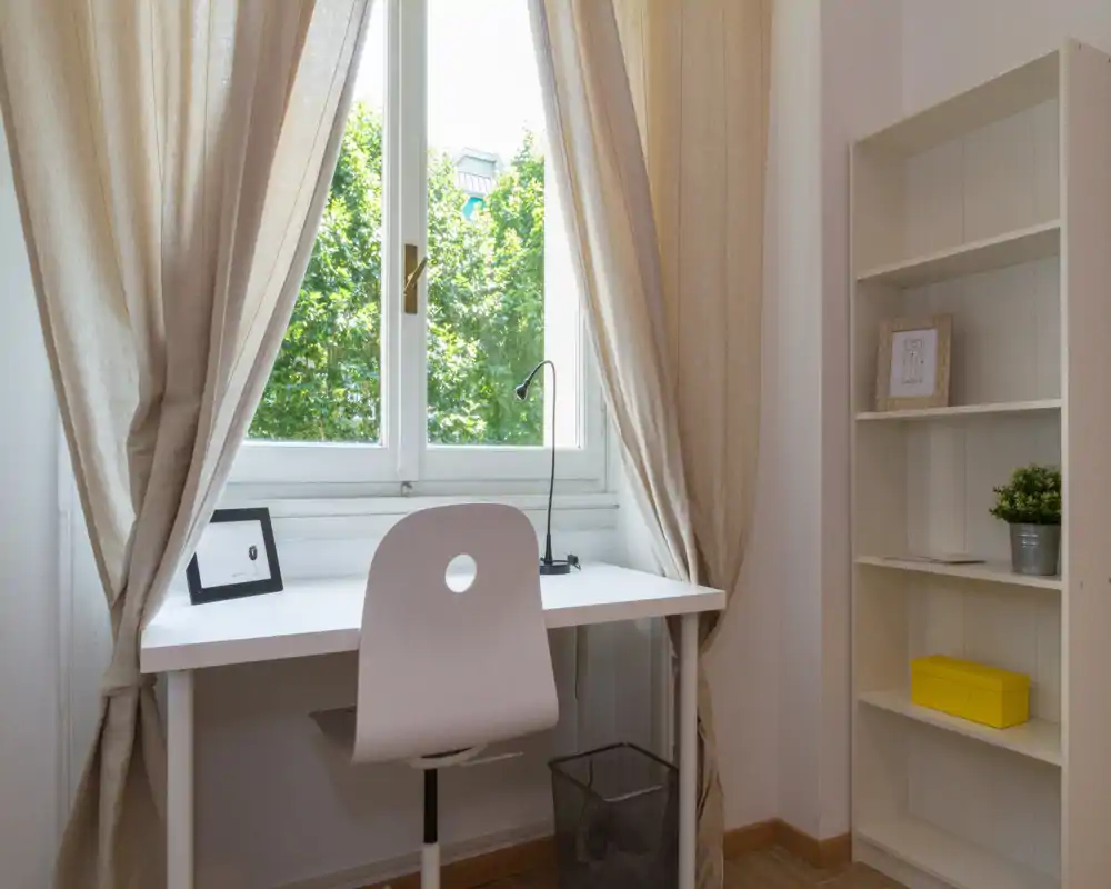 Bright study corner with a white desk and chair positioned at a large window. Soft beige curtains, a slim desk lamp and a small shelving unit create a clean, minimal workspace with natural light.