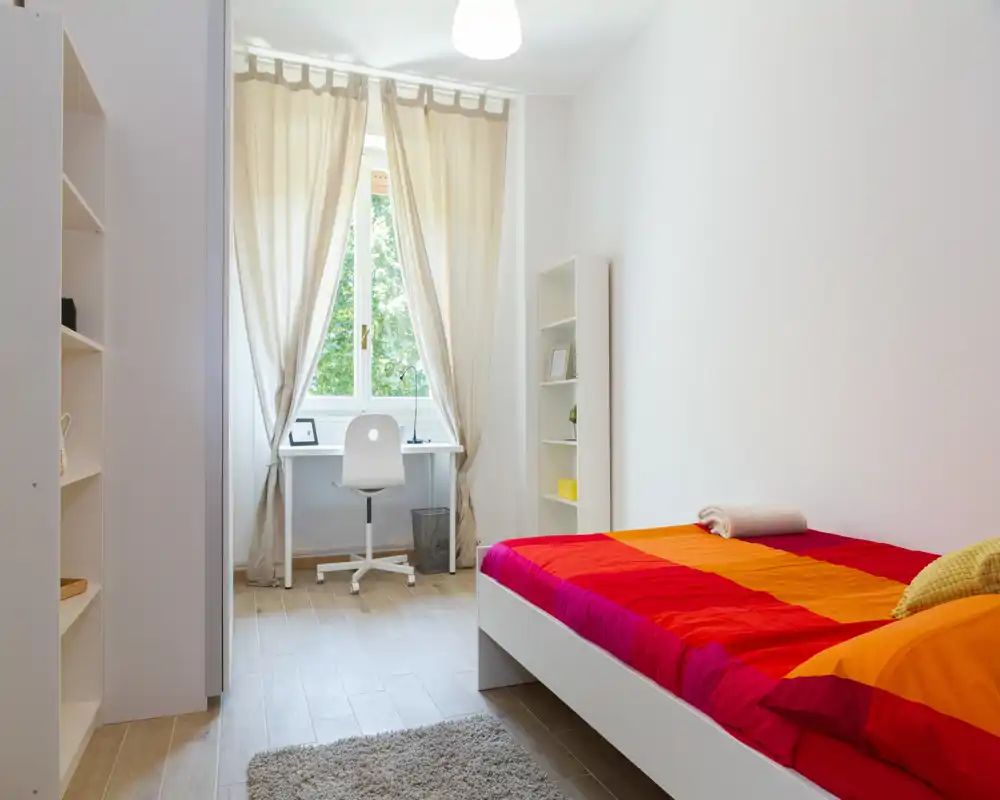 Bright, tidy bedroom with a colorful double bed, wooden floor and a small desk by the window — ideal for relaxing or working. Natural light filters through light curtains creating a warm, welcoming atmosphere.