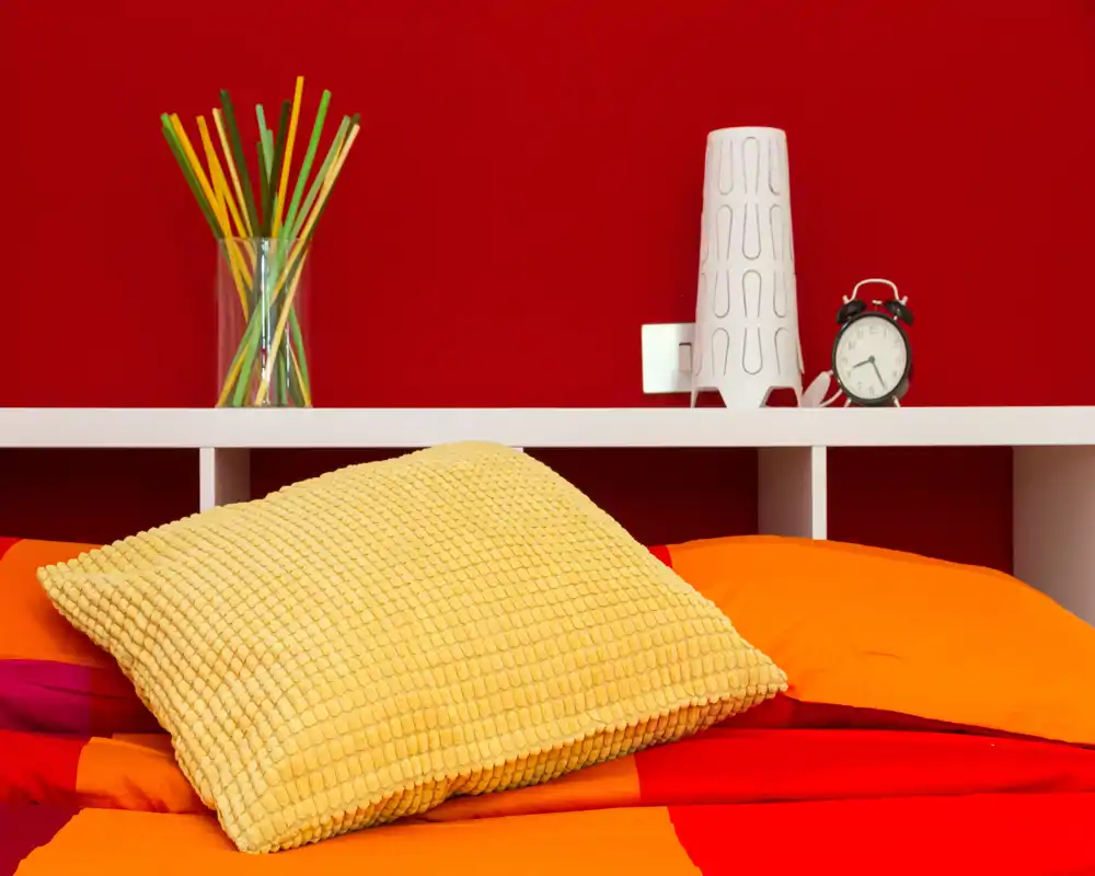 Bright, colorful bedroom close-up with a textured yellow cushion and warm orange-red bedding against a vivid red wall; modern bedside shelf holds a lamp, alarm clock and decorative vase.