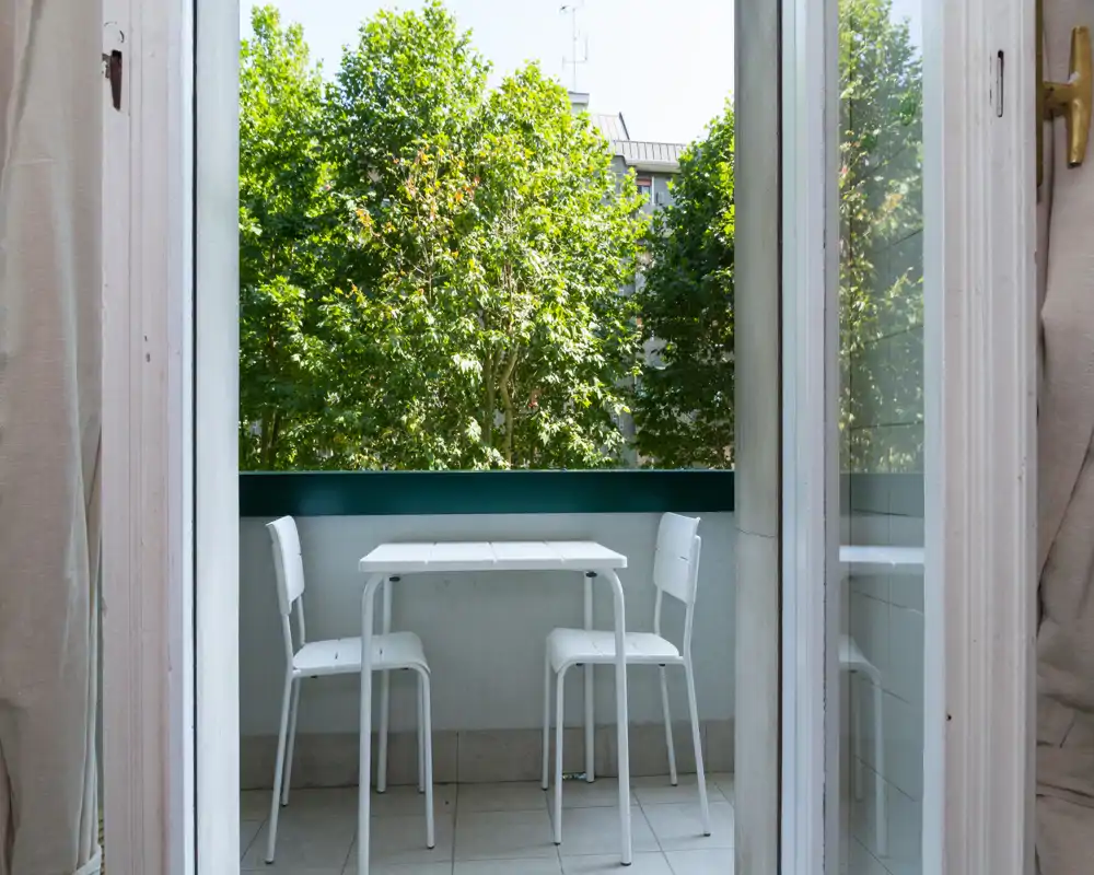 Small private balcony with a compact white table and two chairs, framed by glass doors and leafy green trees beyond — bright and peaceful outdoor spot.