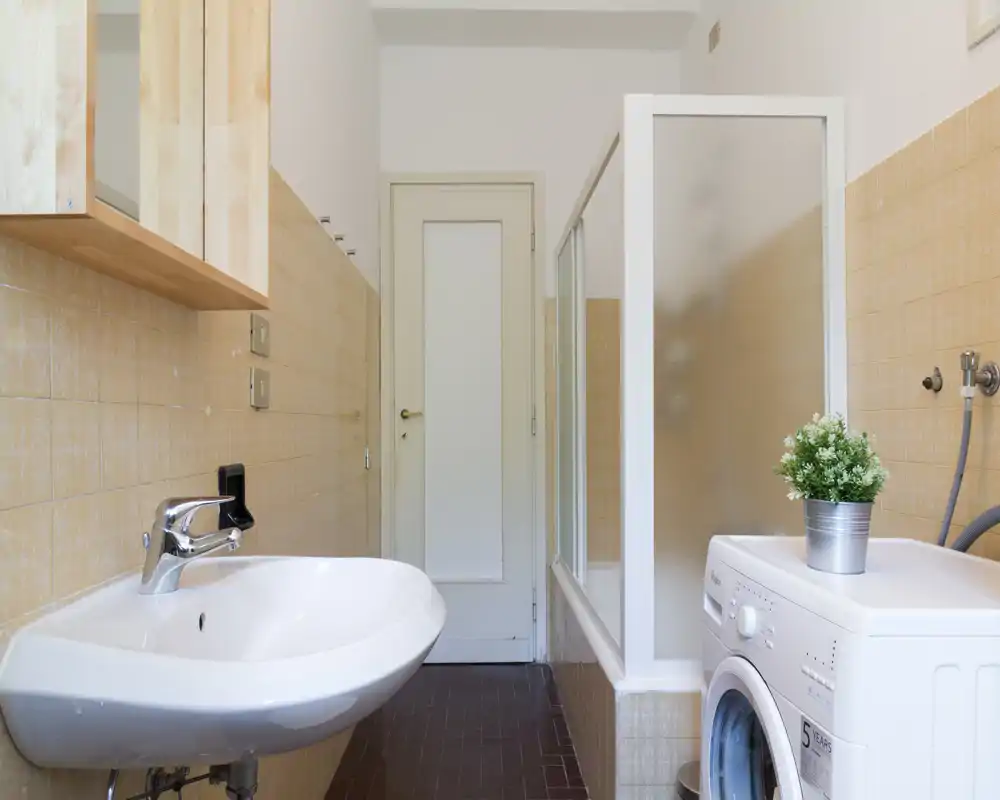 Clean, compact bathroom with a wall-mounted sink, frosted glass shower enclosure and a washing machine; neutral beige tiles and a small plant add a fresh touch.