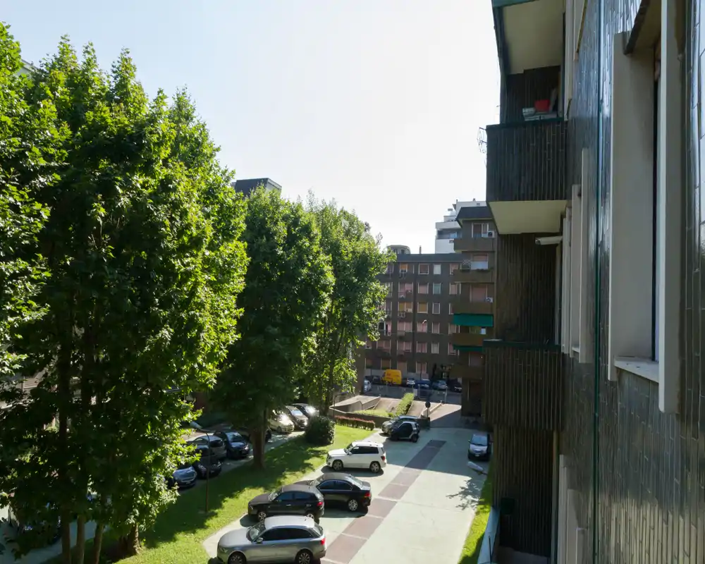 Sunny courtyard view with parked cars, mature trees and apartment building facades — ideal to show communal outdoor parking and green space.