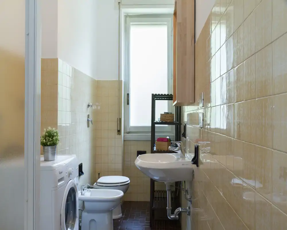 Bright, compact bathroom with a sink, toilet, bidet and washing machine; beige wall tiles and a tall frosted window provide natural light.