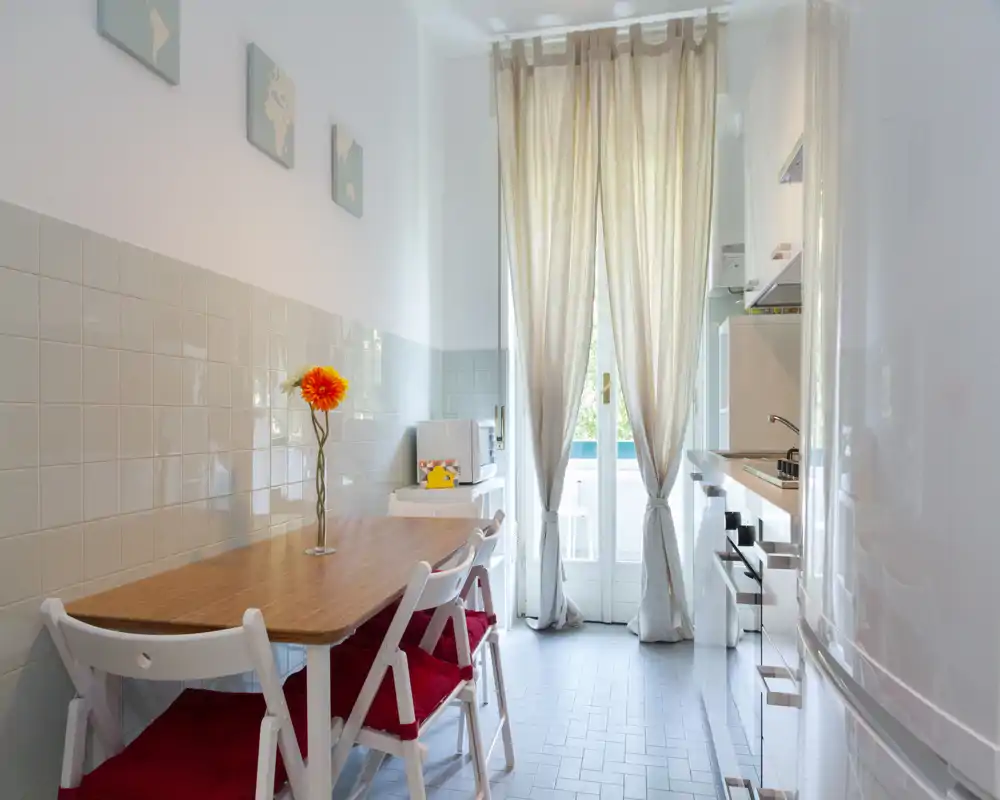 Bright, compact kitchen with a wooden dining table, foldable chairs with red cushions, tiled backsplash and a glass door letting in natural light. The space is neatly styled and suitable for cozy meals.