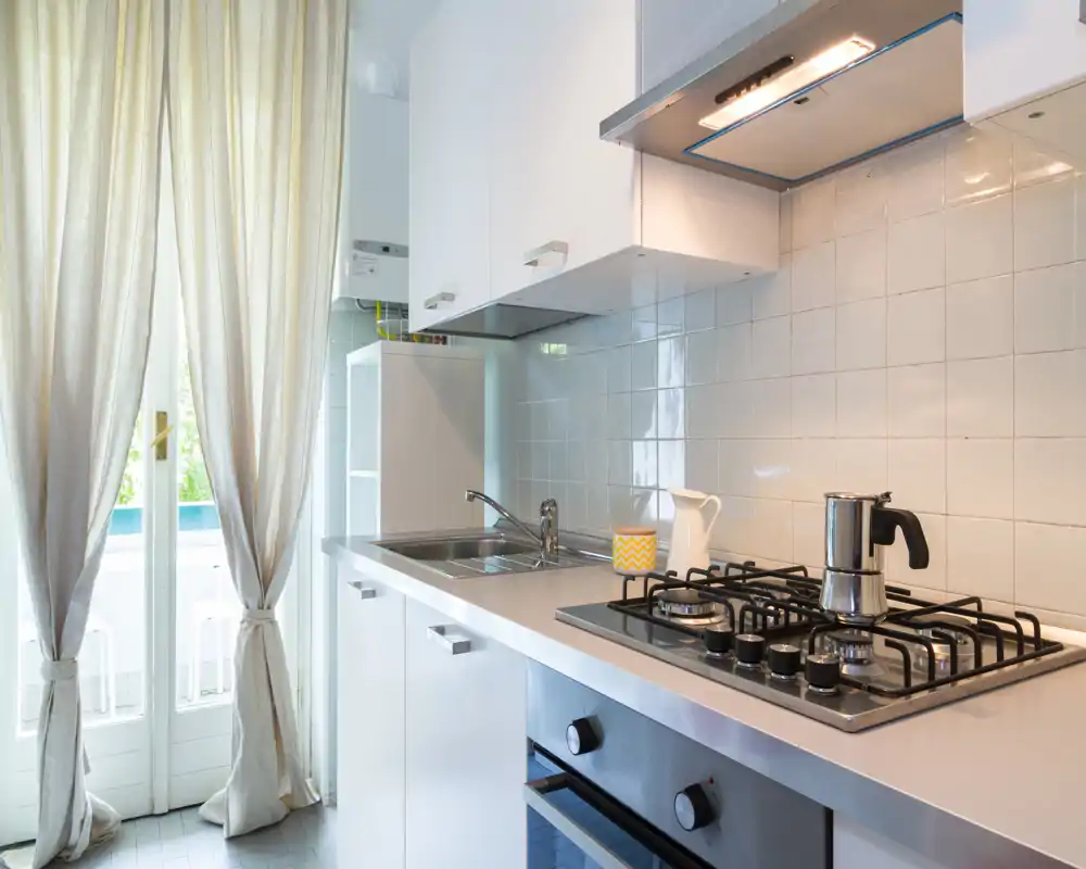 Bright, modern kitchen with a gas hob, stainless sink and sleek white cabinets; natural light enters through full-height glass doors with curtains.