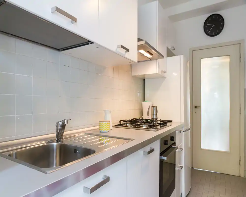 Modern compact kitchen with stainless steel sink, four-burner gas hob and built-in oven. Clean white cabinets, tiled backsplash and pleasant, even lighting.