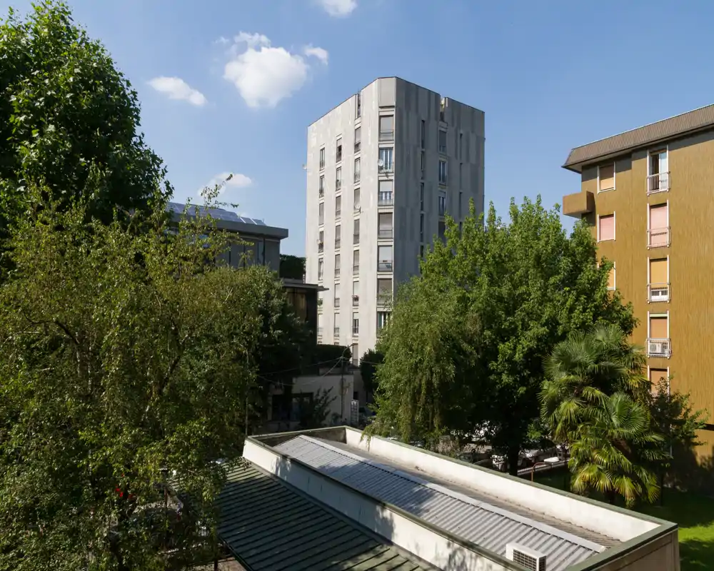 Bright exterior view showing a leafy courtyard with mature trees, nearby residential buildings and a clear blue sky — ideal for listing outdoor space and neighborhood views.