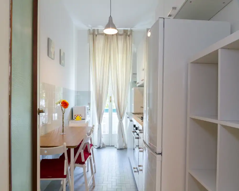 Bright narrow kitchen with a small wooden dining table and red-cushioned chairs, white cabinets and a large window with sheer curtains that fill the space with natural light.