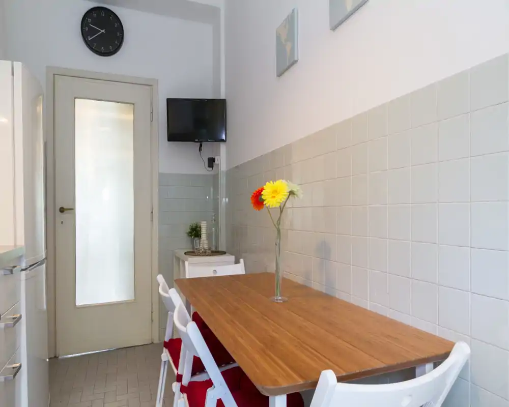 Bright, tidy kitchen dining area with a wooden table, white folding chairs and a simple floral centerpiece. Light gray tiled walls and a small wall-mounted TV complete the compact, functional space.