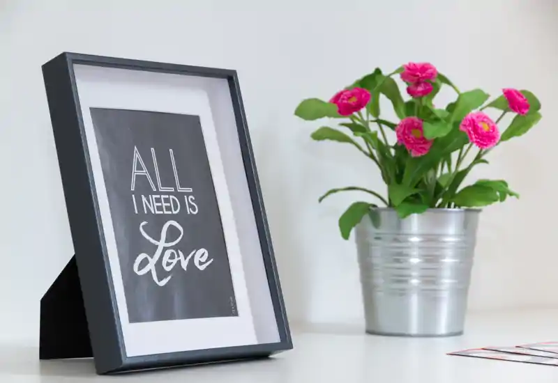 Scatto ravvicinato di decor: una cornice con la scritta “All I need is Love” accanto a un vaso metallico con fiori rosa su una superficie bianca — perfetto per mostrare dettagli decorativi o elementi di styling.