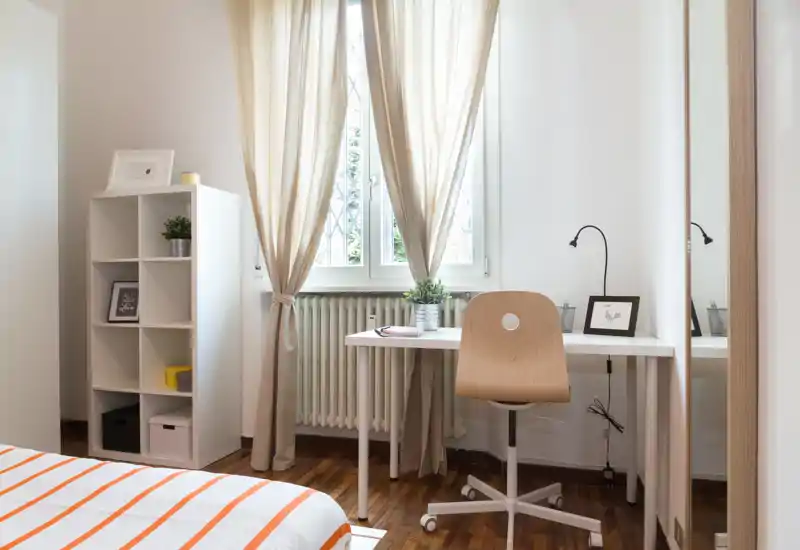 Angolo camera da letto luminoso e ordinato con letto a righe in primo piano, scrivania davanti alla finestra e libreria per riporre oggetti. La luce naturale, le tende calde e il pavimento in legno creano un’atmosfera accogliente, perfetta per annunci.