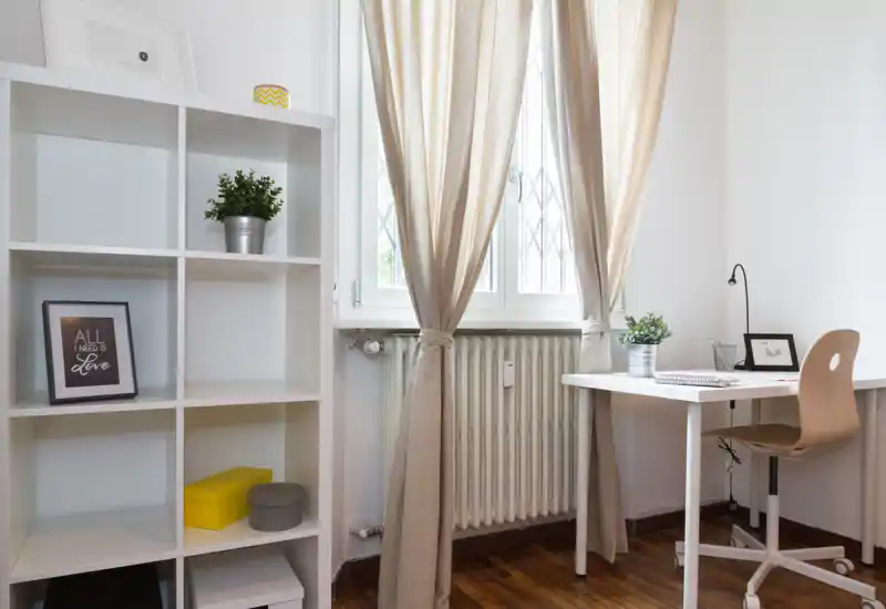 Angolo di camera luminoso e compatto con scrivania, libreria a cubi e finestra con tende chiare; stile minimal e luce naturale rendono l’ambiente accogliente.