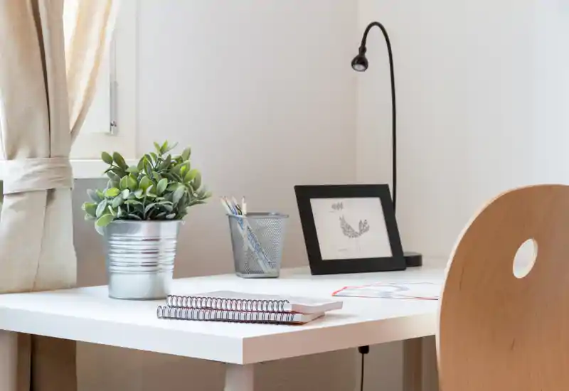 Angolo scrivania luminoso e minimal con tavolo bianco, sedia in legno, pianta in vaso e lampada da lavoro — ideale per un piccolo ufficio domestico o zona studio.