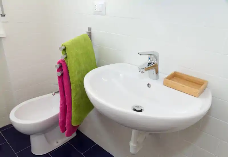 Bagno moderno e compatto con lavabo a parete, rubinetto cromato, bidet e asciugamani colorati su un piccolo portasciugamani; piastrelle bianche e pavimento scuro per un aspetto pulito e contemporaneo.