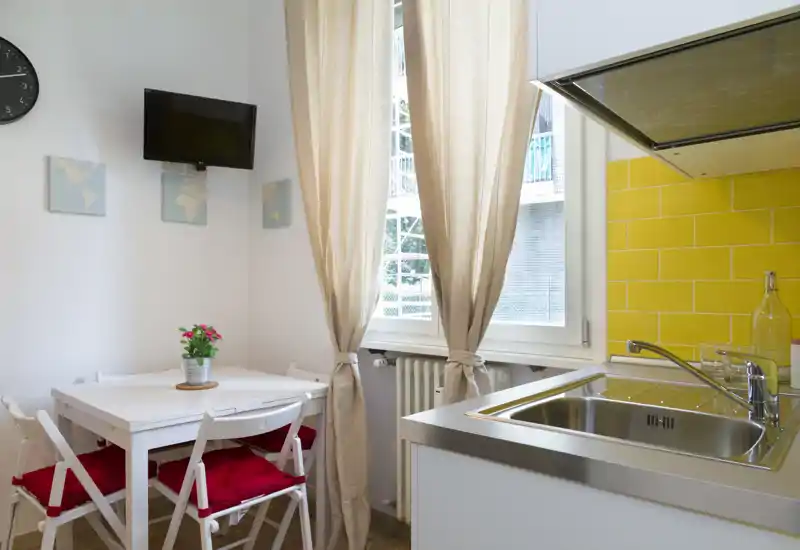 Angolo cucina moderno e compatto con lavello in acciaio, schienale a piastrelle gialle e un piccolo tavolo da pranzo vicino alla finestra luminosa—accogliente e ben illuminato.