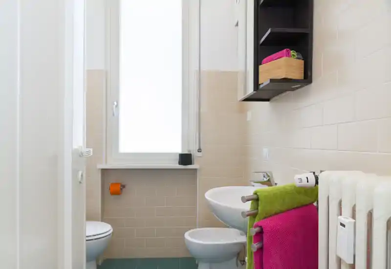 Bagno moderno e luminoso con lavabo a parete, bidet, WC e asciugamani colorati; piastrelle neutre e una grande finestra smerigliata che diffonde luce naturale.