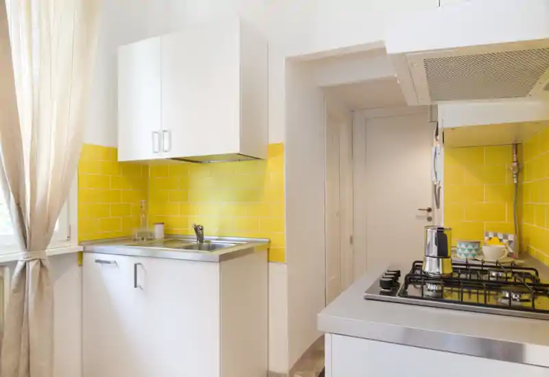 Cucina moderna e compatta con paraschizzi in piastrelle gialle, lavello in acciaio inox e piano cottura a gas. Luce naturale dalla finestra e pensili bianchi puliti rendono l'ambiente fresco e luminoso.