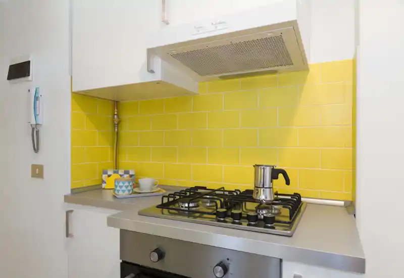 Angolo cucina moderno e compatto con piano cottura a gas in acciaio, cappa aspirante e retrocucina con piastrelle gialle; tazze e una moka aggiungono carattere.