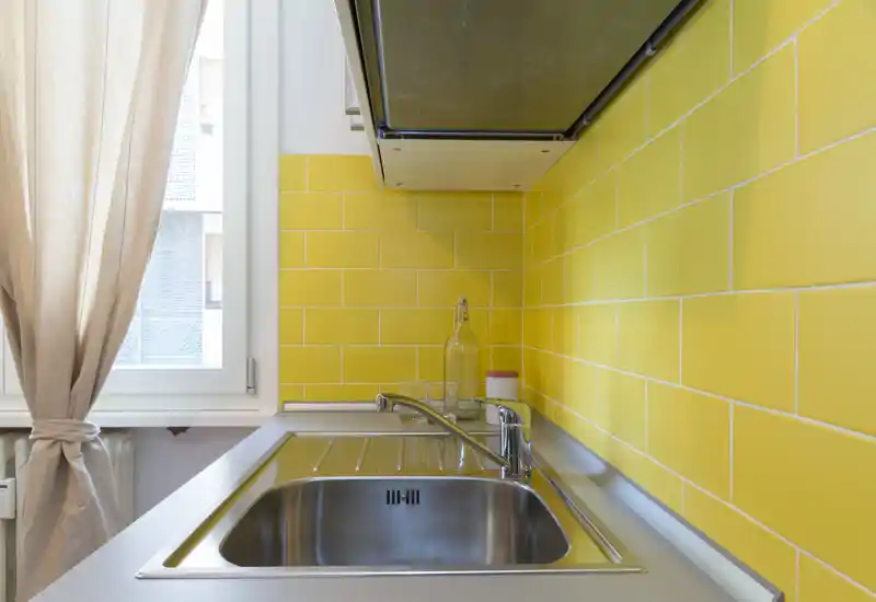 Zona lavello cucina compatta e luminosa con vasca in acciaio inox lucido, miscelatore moderno e vivace retrostante in piastrelle gialle vicino a una finestra.