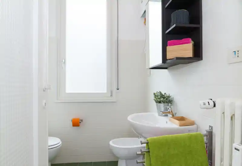 Bagno compatto e luminoso con lavabo bianco, bidet, WC e finestra satinata; asciugamano verde e mensola con accessori per praticità.