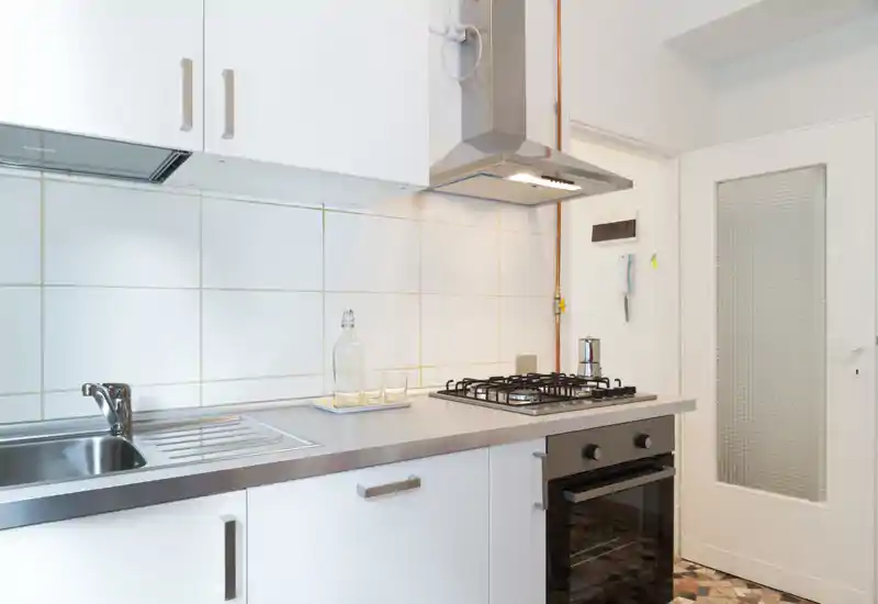 Cucina moderna e luminosa con piano in acciaio, lavello integrato, piano cottura a gas a quattro fuochi e cappa. Pensili bianchi e parete piastrellata offrono un aspetto pulito e minimale, perfetto per annunci.