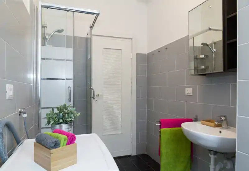 Bagno moderno e pulito con box doccia angolare, lavabo a parete, specchiera e asciugamani colorati che donano un aspetto fresco.