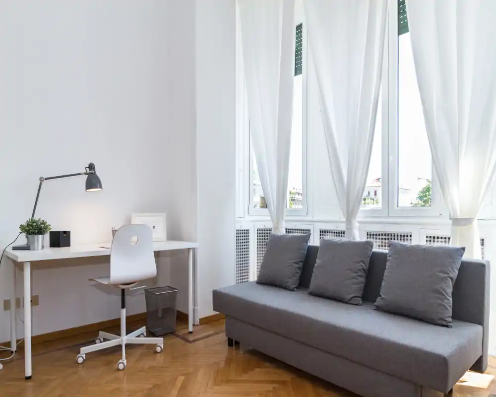 Zona de estar luminosa y minimalista con sofá gris y tres cojines, un escritorio con silla junto a grandes ventanas que aportan mucha luz natural — ideal para descansar o trabajar.