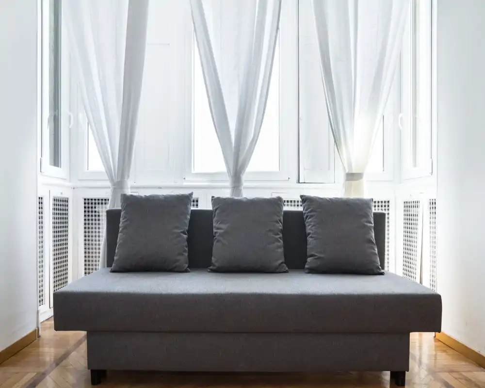 Zona de estar luminosa y minimalista con un sofá gris centrado y tres cojines frente a grandes ventanas con cortinas translúcidas.