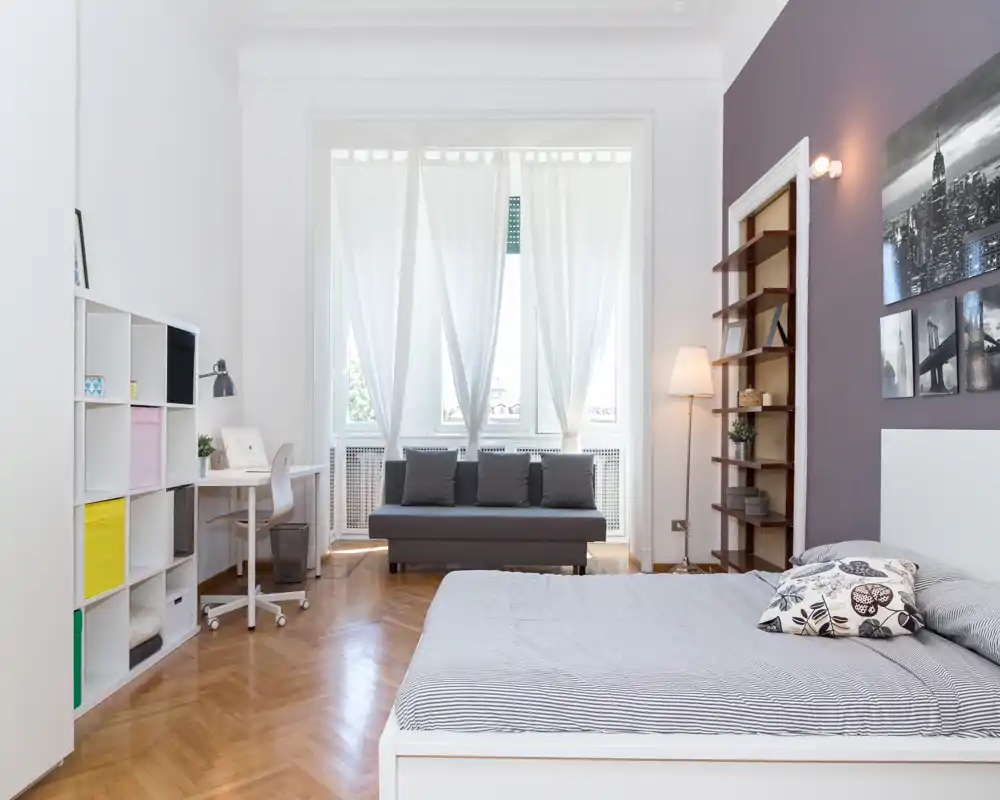 Dormitorio amplio y luminoso con cama en primer plano, zona de estar acogedora junto a una gran ventana y estanterías funcionales — perfecto para una estancia cómoda.