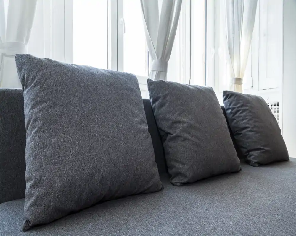 Primer plano de un sofá gris moderno con tres grandes cojines frente a ventanas luminosas, mostrando la textura de la tela y un ambiente neutral y tranquilo.