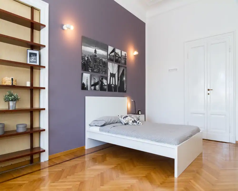 Dormitorio luminoso y minimalista con cama blanca frente a una pared color púrpura suave, fotos en blanco y negro, lámpara de mesita y suelo de parqué.