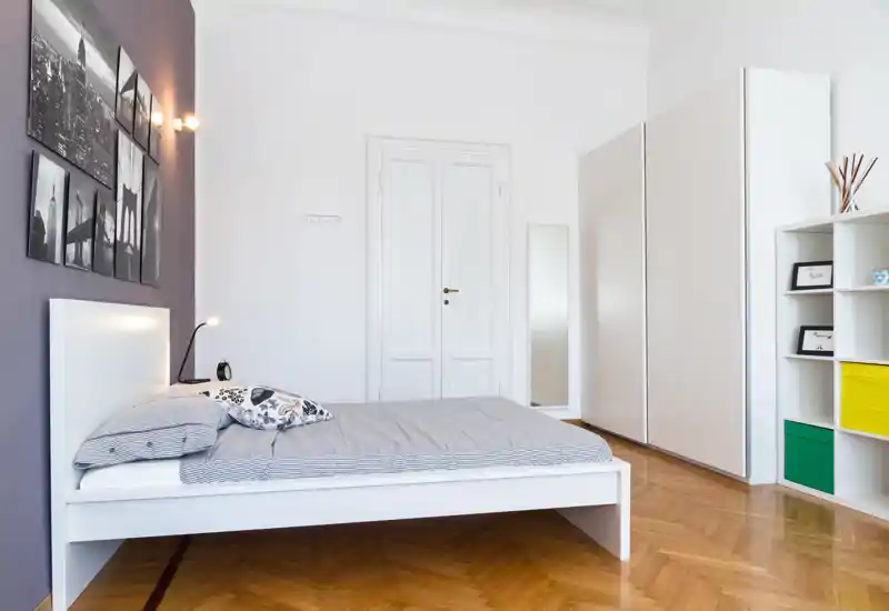 Dormitorio moderno y luminoso con cama blanca baja, ropa de cama a rayas y cojines decorativos; gran armario y estantería que ofrecen amplio almacenaje. Suelo de parquet limpio y arte mural minimalista crean un ambiente tranquilo y con estilo, ideal para anuncios.