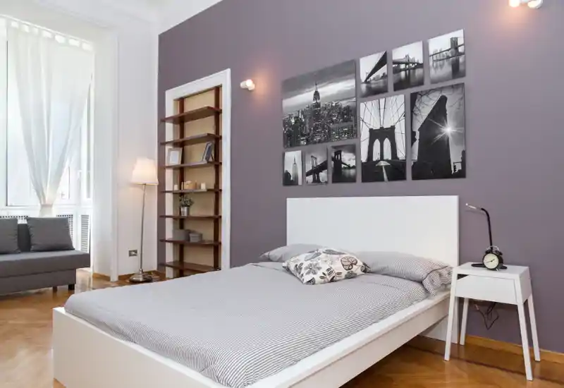 Dormitorio moderno y luminoso con cama blanca de plataforma, cojines decorativos y cuadros en blanco y negro sobre la cabecera; gran ventana que aporta luz natural y suelos de madera que aportan calidez.