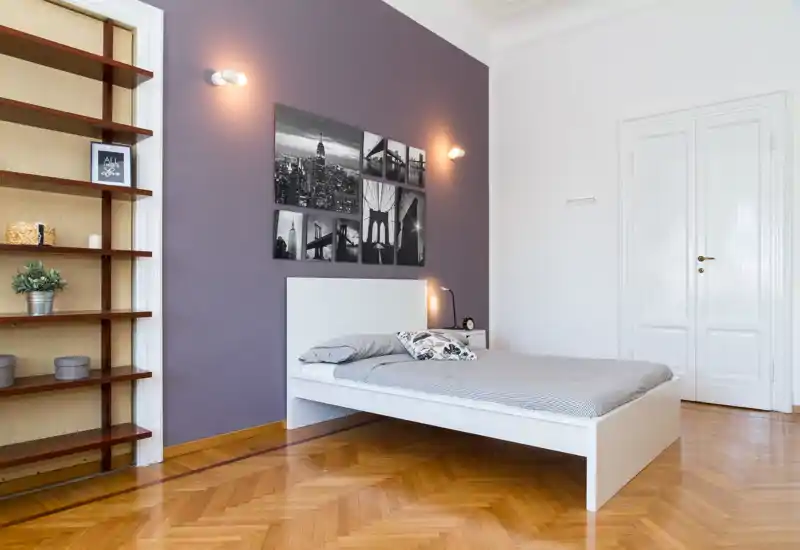 Dormitorio luminoso y minimalista con cama blanca frente a una pared color púrpura suave, fotos en blanco y negro, lámpara de mesita y suelo de parqué.