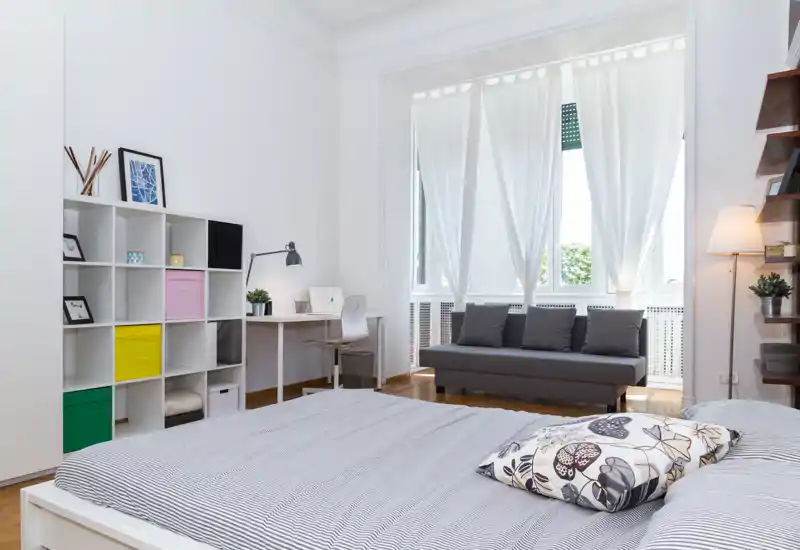 Dormitorio luminoso y con estilo con la cama bien hecha en primer plano, un pequeño escritorio y estanterías a la izquierda y un sofá junto a una gran ventana que aporta mucha luz natural.