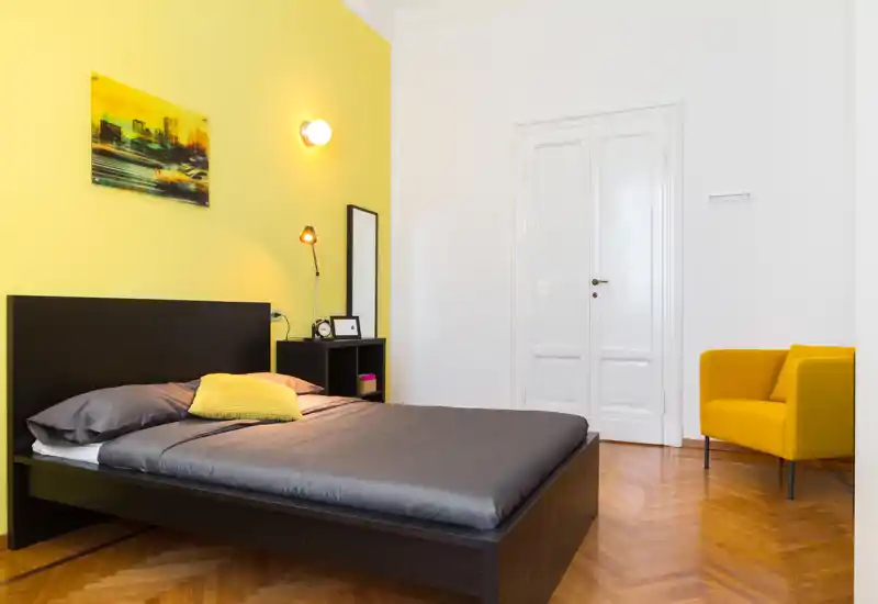 Bright, modern bedroom with a dark wood bedframe, gray bedding and a yellow accent wall. The room features a bedside shelf, floor lamp and a yellow armchair, creating a warm, stylish feel ideal for rental photos.