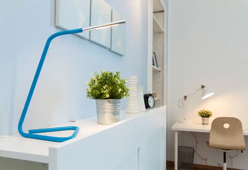 Rincón de trabajo luminoso y minimalista con una lámpara de escritorio azul moderna, planta en maceta y silla — perfecto como espacio de estudio u oficina en casa.