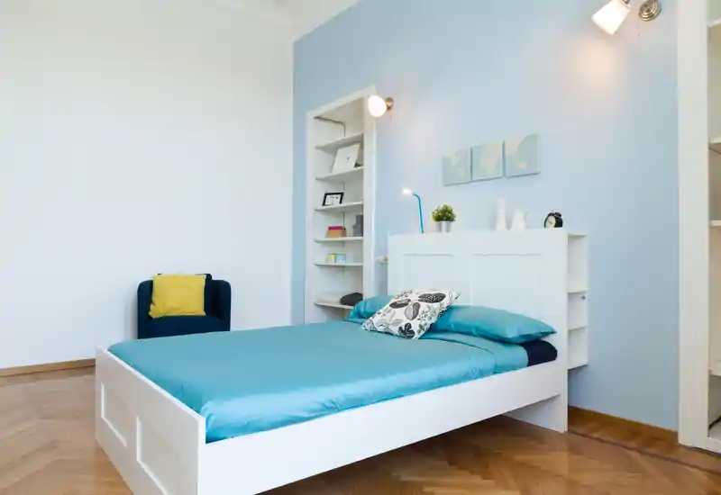 Dormitorio luminoso y bien presentado con estructura de cama blanca y ropa de cama turquesa, suelo de parquet y un sillón en la esquina.