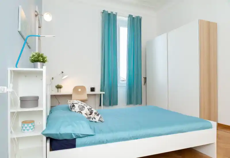 Dormitorio moderno y luminoso con cama doble bien hecha en ropa de cama turquesa, armario blanco y un pequeño escritorio junto a la ventana — ideal para una estancia cómoda.