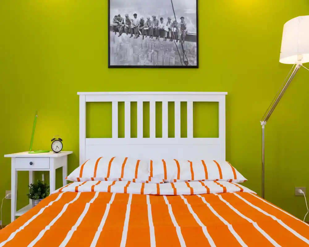 Bright, modern bedroom with a white slatted headboard and an orange-and-white striped duvet. The room features a bedside table with alarm clock and a tall floor lamp, set against a vivid green accent wall.