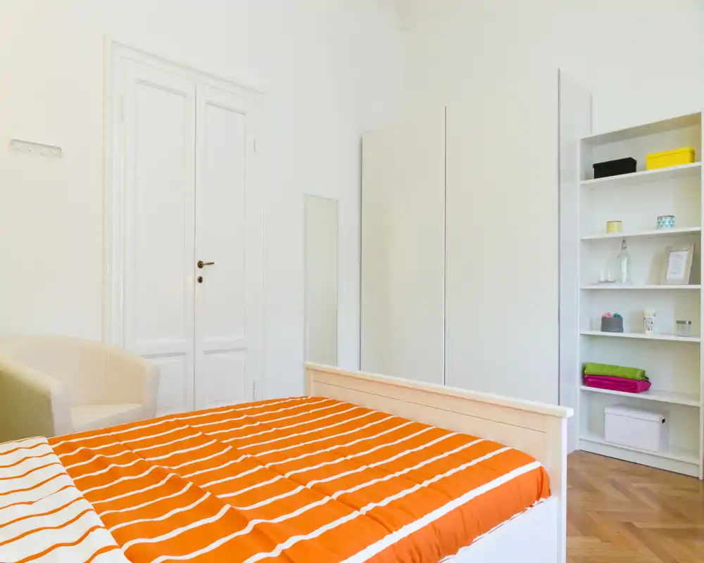 Bright, tidy bedroom with a white bed frame and orange-striped bedding. The room features a wardrobe, shelving and a small armchair, creating a simple, welcoming space.